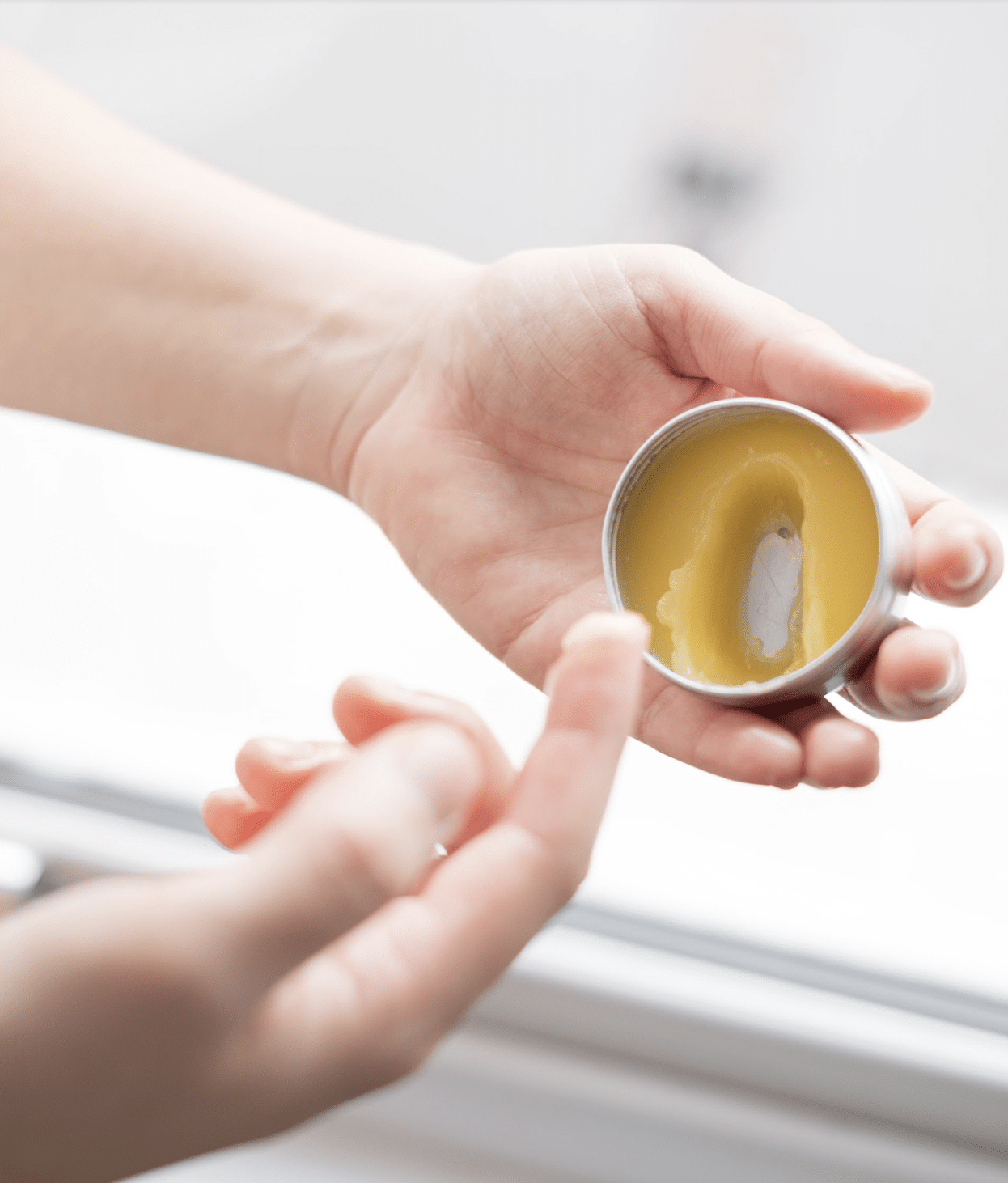 Image is a pair of hands reaching into a jar of moisturizer. The jar is silver and the balm is a yellow, natural color.