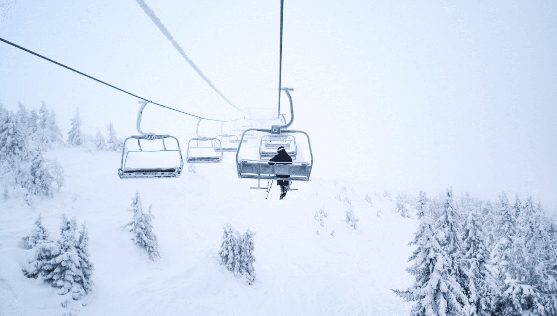 Image of a ski lift in the snow. There is one person riding and looking out over the snow covered treetops.