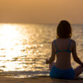 Image of the back of a woman overlooking a sunrise sitting on a yoga mat ready to start the day with meditation.