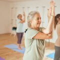 A class of older women practicing yoga. They all have their hands clasped together as they stand on blue mats.