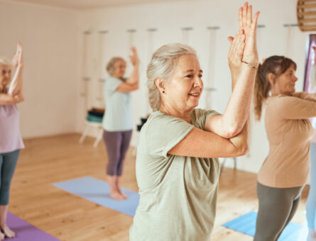 A class of older women practicing yoga. They all have their hands clasped together as they stand on blue mats.